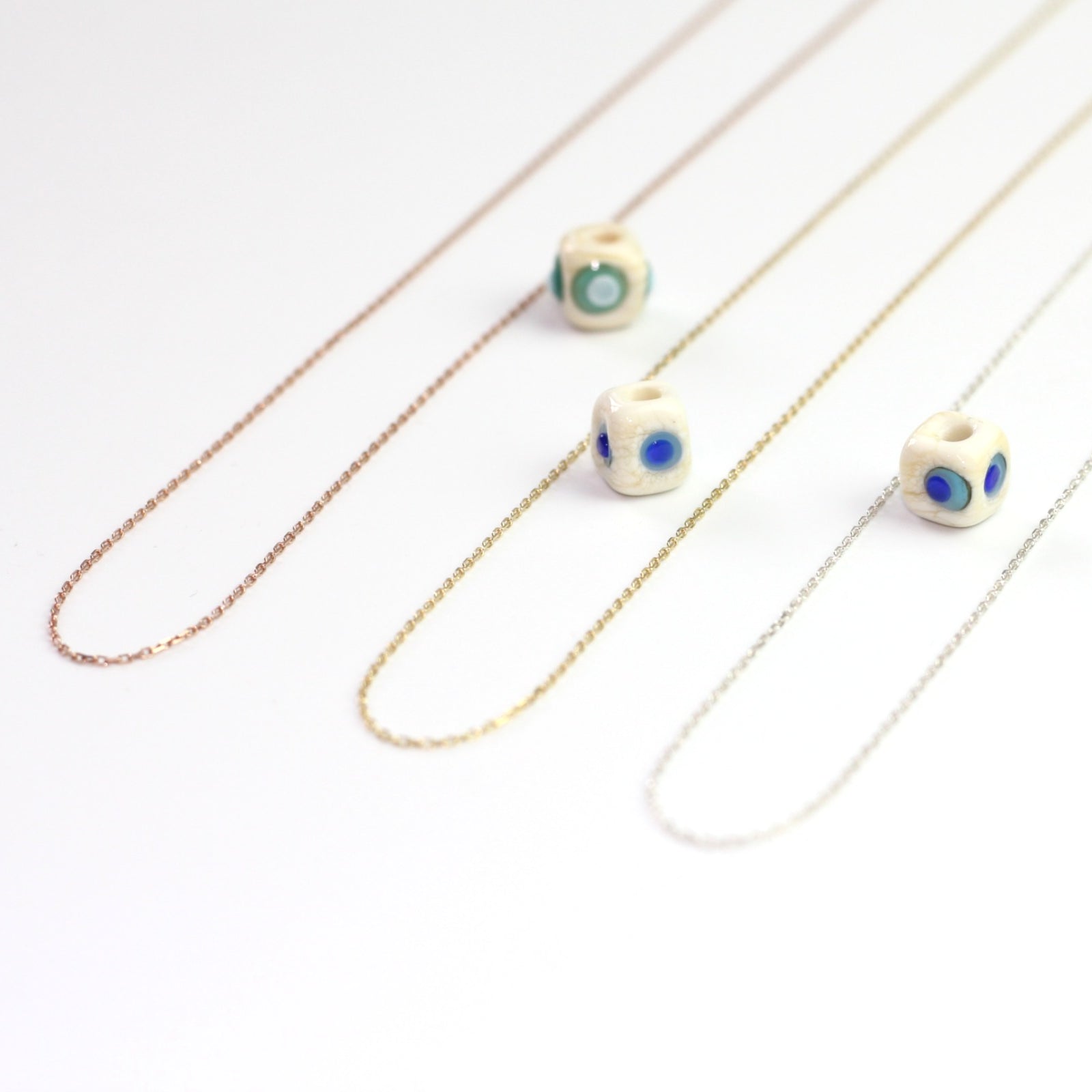 Yakın çekim, küp şeklinde el yapımı cam nazar boncuğu kolye ucu. Kolye ucu cam sanatı ürünüdür ve 0,5 köşe uzunluğundadır. Ürün, ortasından geçen 50 cm gümüş, altın veya rose rengi zincirle gösteriliyor.