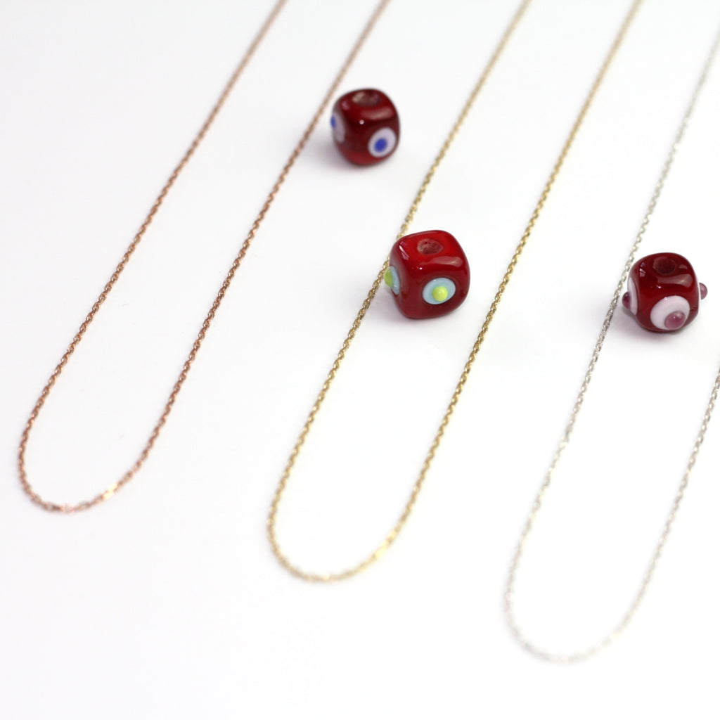 Yakın çekim, küp şeklinde el yapımı cam nazar boncuğu kolye ucu. Kolye ucu cam sanatı ürünüdür ve 0,5 köşe uzunluğundadır. Ürün, ortasından geçen 50 cm gümüş, altın veya rose rengi zincirle gösteriliyor.