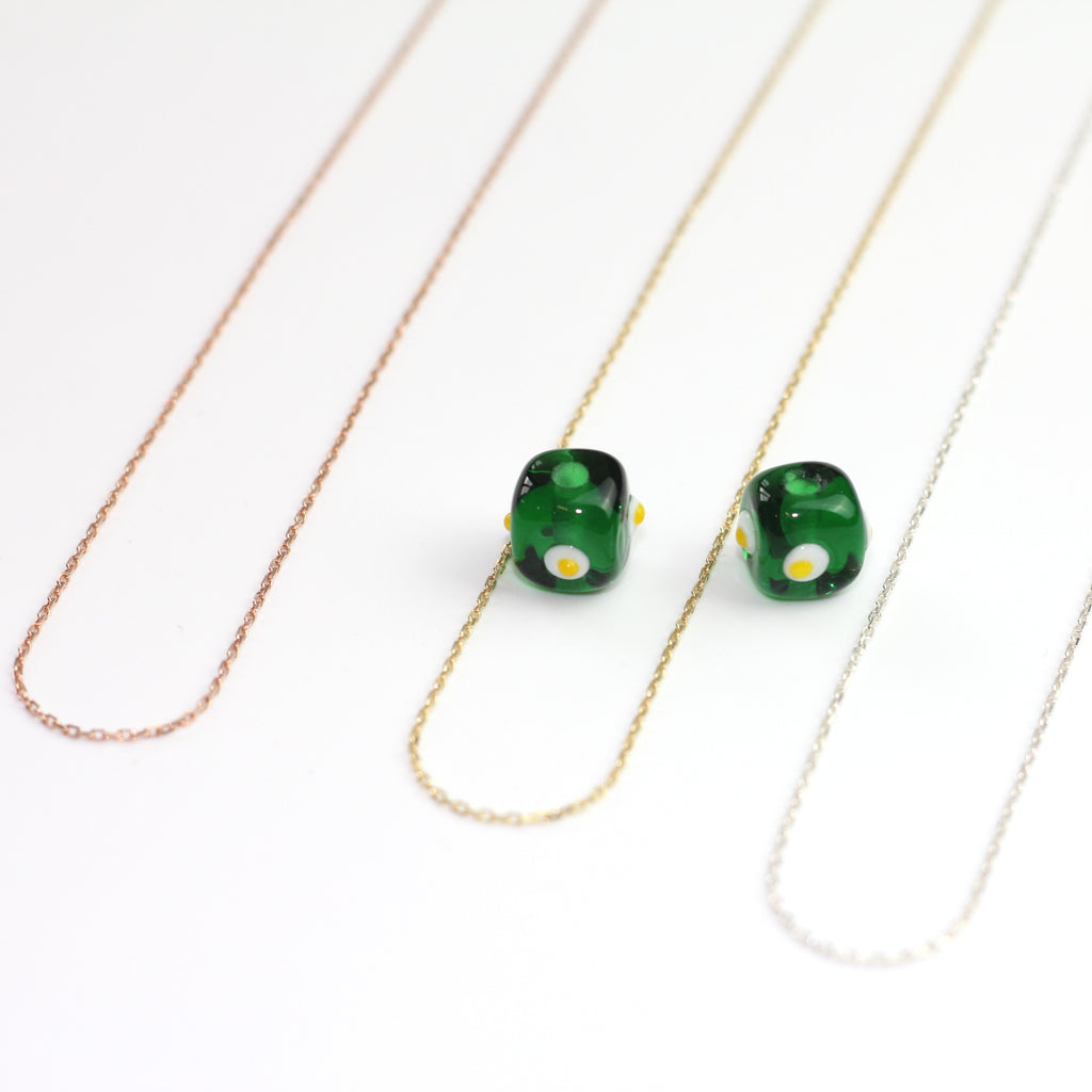 Yakın çekim, küp şeklinde el yapımı cam nazar boncuğu kolye ucu. Kolye ucu cam sanatı ürünüdür ve 0,5 köşe uzunluğundadır. Ürün, ortasından geçen 50 cm gümüş, altın veya rose rengi zincirle gösteriliyor.